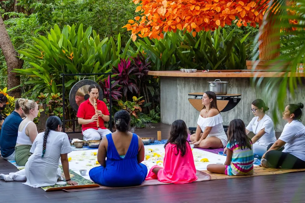 A group of people seated around an instructor giving a meditation class in a serene, tree-filled natural place. The instructor is playing a large wind instrument similar to a clarinet, creating a relaxing atmosphere.